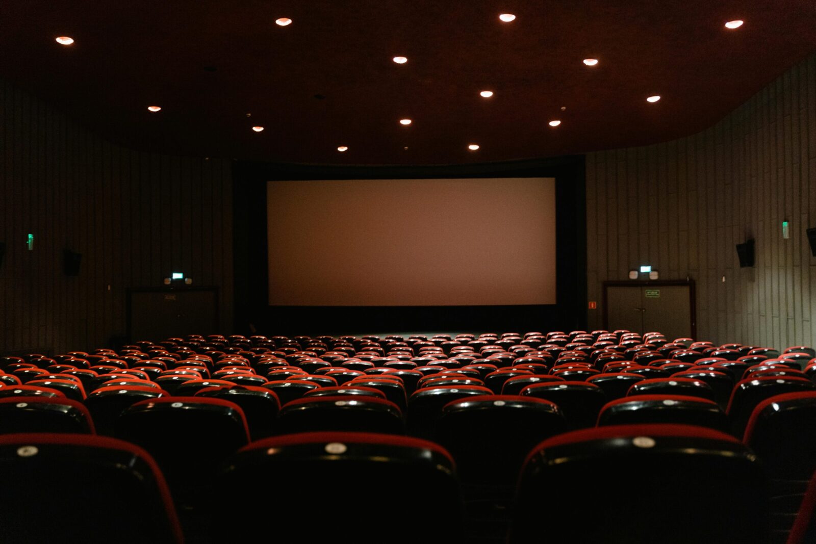 A dimly lit empty cinema theater with rows of red chairs facing a projector screen.